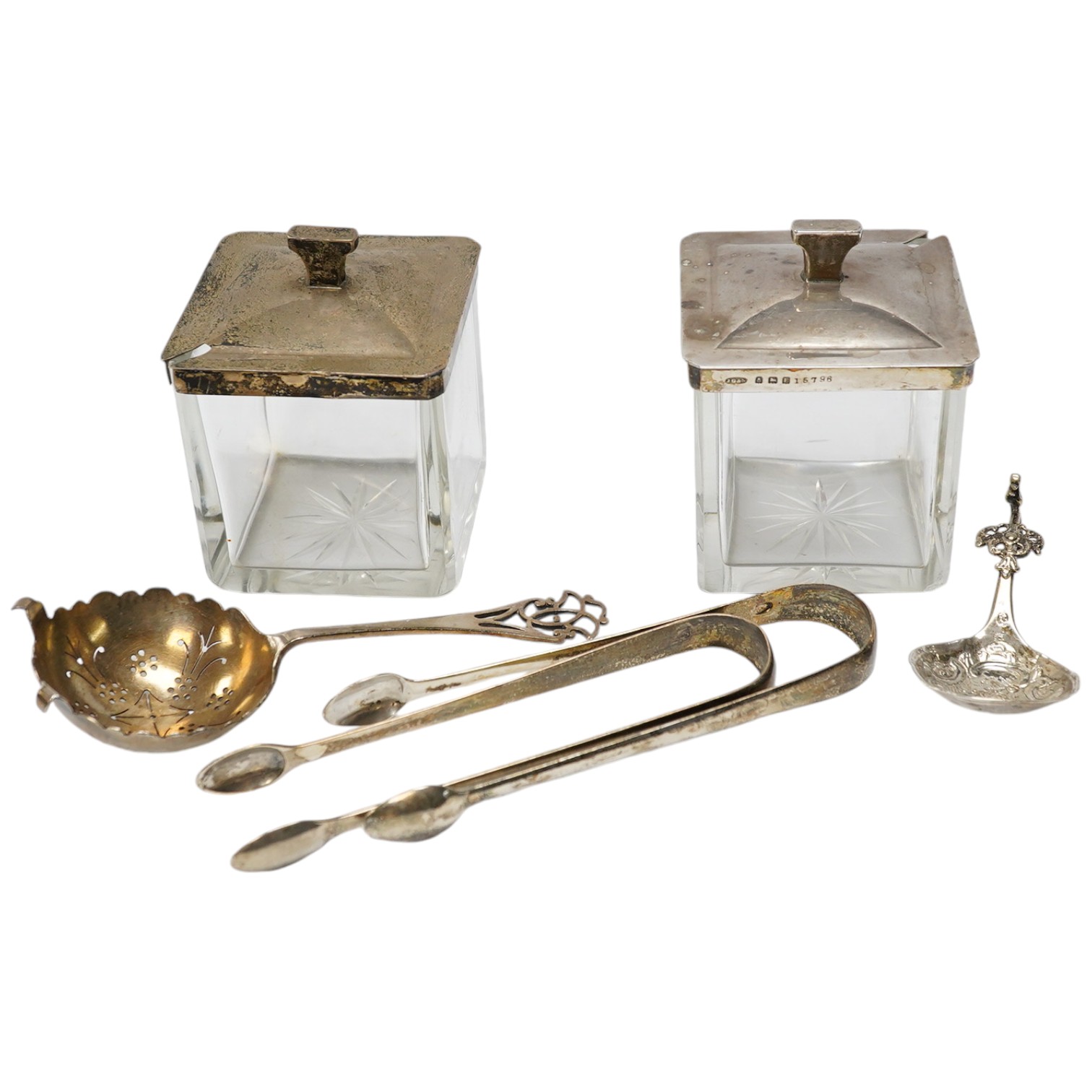 Two square silver topped glass preserve pot, two pairs of George III silver sugar tongs, a silver tea strainer and a caddy spoon. Condition - fair